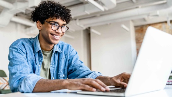 Junger Mann mit Brille sitzt lachend vor einem Laptop