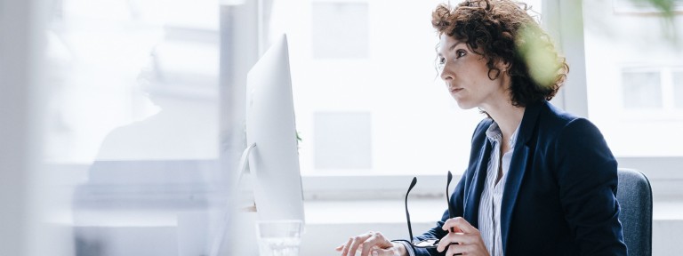 Frau hält ihre Brille in der Hand und schaut in einen Monitor auf ihrem Schreibtisch