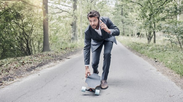 Mann im Anzug steht auf einem Skateboard und telefoniert dabei mit seinem Handy