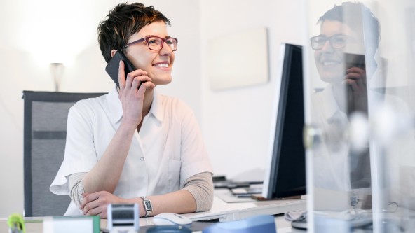 Frau mit roter Brille sitzt telefonierend vor einem Monitor