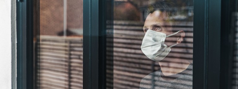 Mann steht mit weißem Mundschutz vor einem Fenster und schaut nach draußen