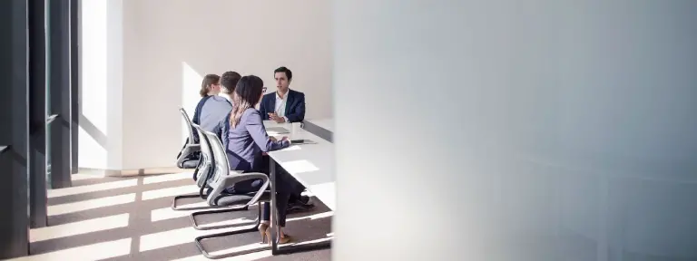Neben einer Säule sitzen im Hintergrund 4 Personen in einem Besprechungsraum