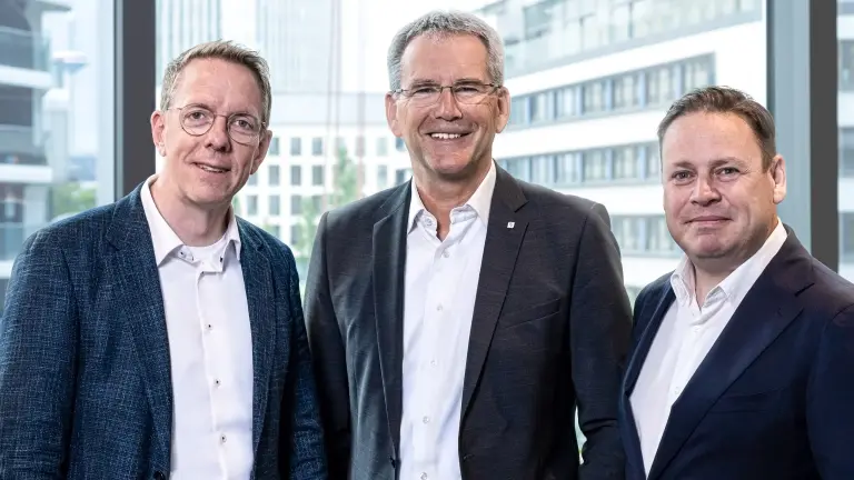 Drei Männer in Anzügen stehen vor einer Fensterfront