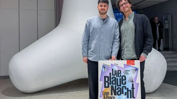 Zwei Personen stehen mit einem Werbeschild für die Blaue Nacht vor einer grauen Kunstinstallation
