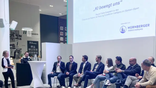 9 Personen sitzen für ein Rounttable-Gespräch nebeneinander, zwei Moderatoren sind ebenfalls auf der Bühne