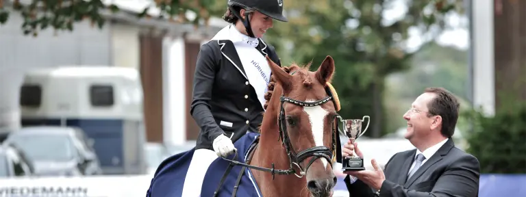Junge Frau sitz auf einem Pferd und bekommt von einem Mann einen Pokal überreicht